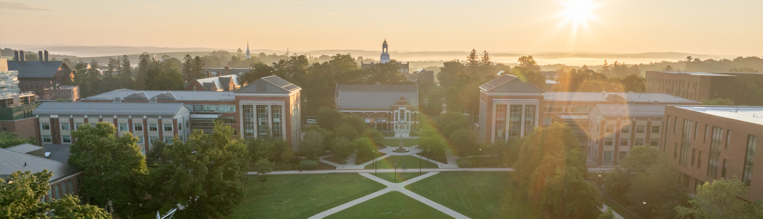 Aerial view of a UConn campus, a large green lawn in the foreground with buildings in the background, on a hazy summer day.